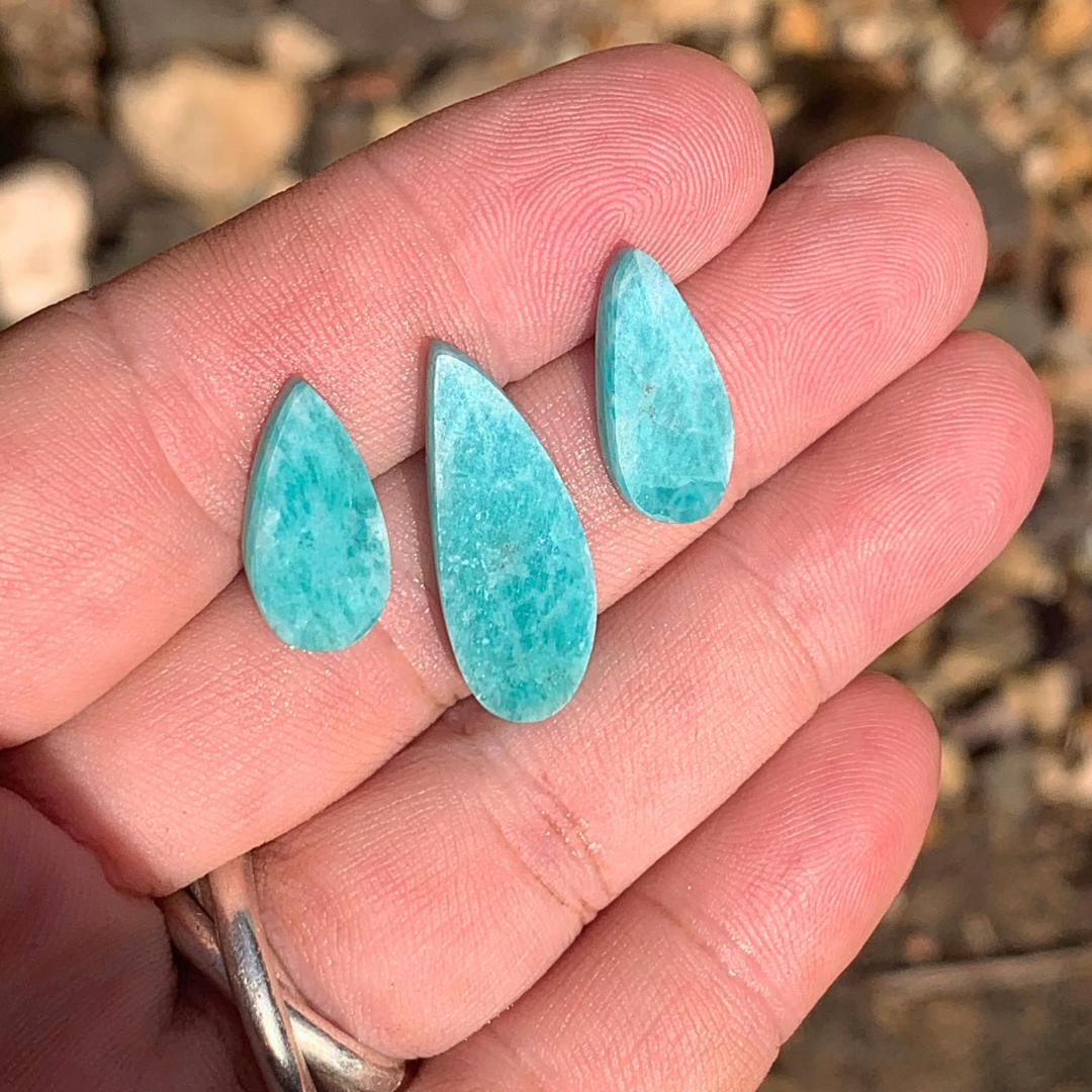 Three teardrop-shaped turquoise blue amazonite gemstones held between fingers against a blurred natural background in ths sunshine