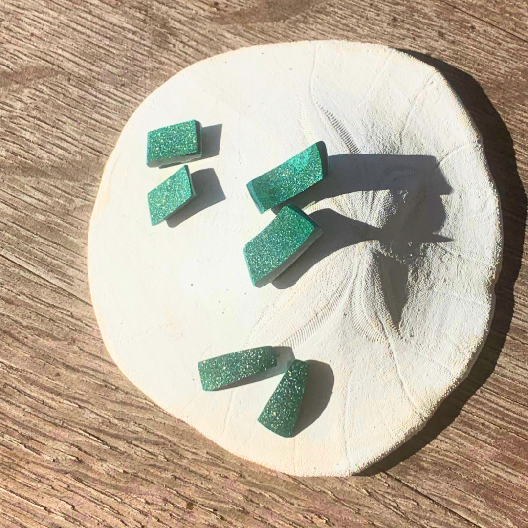 Ray of Sunshine Jewellery- Green glittery gemstone pairs on a white sand dollar shell with a wooden background in the sunshine