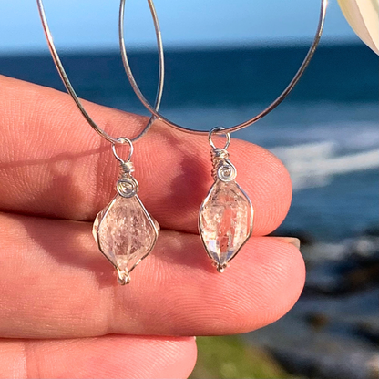Ray of Sunshine Jewellery- Pair of silver hoop earrings with Herkiner Diamond gemstone drops held against a blurred beach background.