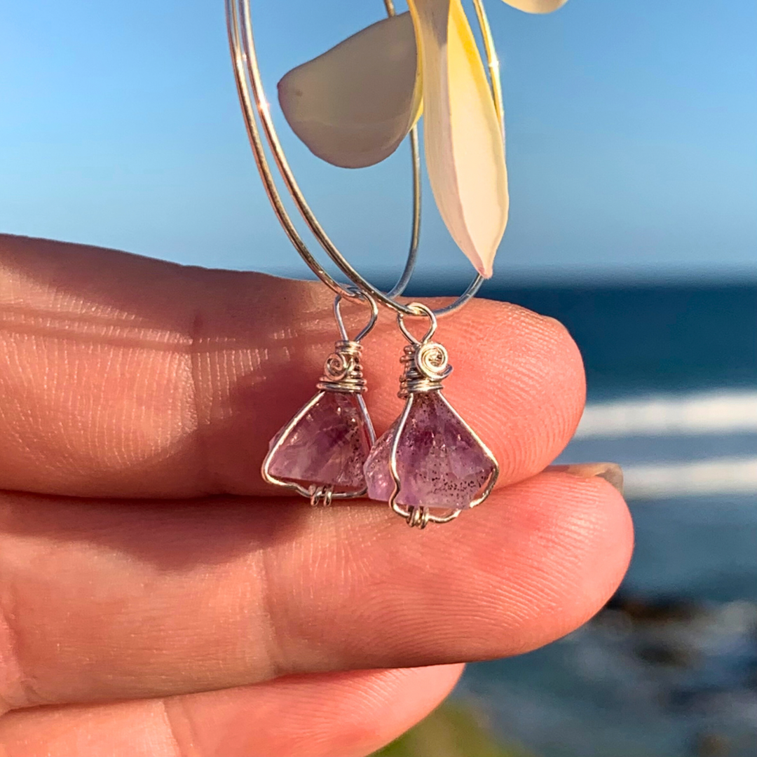 Ray of Sunshine Jewellery- Amethyst earrings held between fingers in the sunshine with a blurred beach background