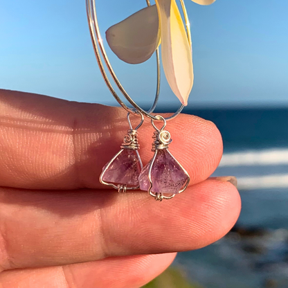 Ray of Sunshine Jewellery- Amethyst earrings held between fingers in the sunshine with a blurred beach background