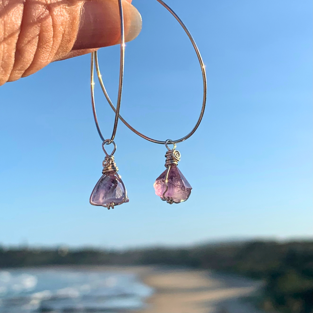 Ray of Sunshine Jewellery- Purple Amethyst point gemstone earrings held up against a blue sky with a blurred beach background