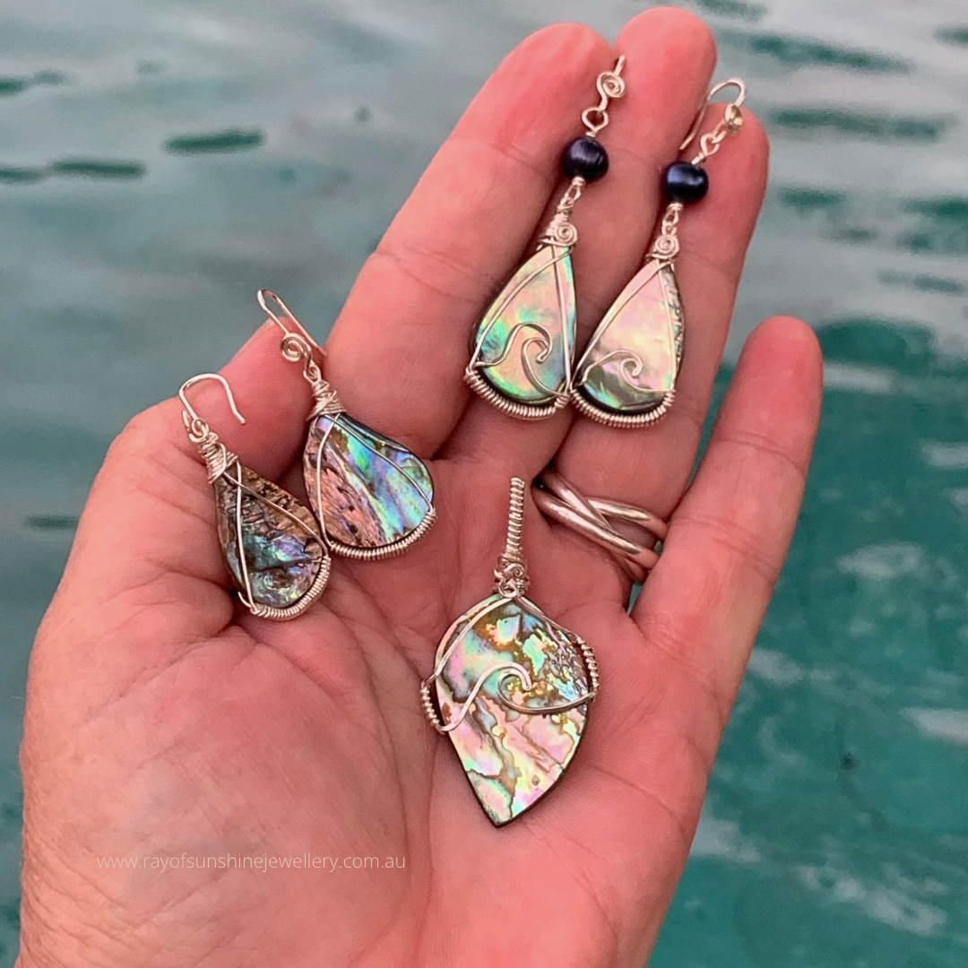 Hand holding three teardrop-shaped multicoloured shell jewelry pieces with a water background
