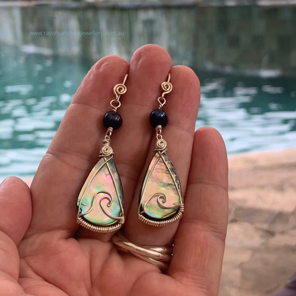 Multicoloured teardrop-shaped albalone earrings with silver wire wrapping held in my hand overlooking shimmering water.