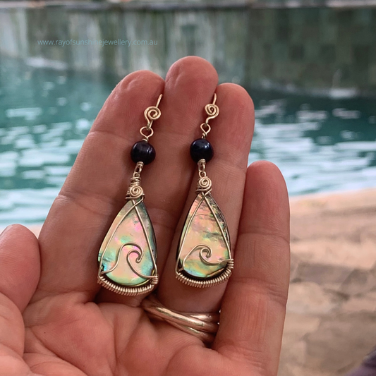 Multicoloured teardrop-shaped albalone earrings with silver wire wrapping held in my hand overlooking shimmering water.