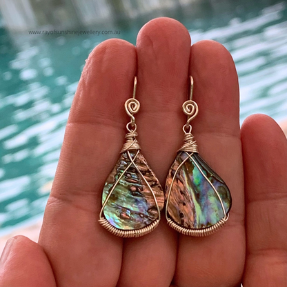 Multicolored albalone shell teardrop-shaped earrings with silver wire wrapping held between fingers against a blurred water background.