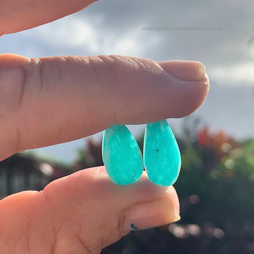 Turquoise translucent amazonite teardrop gemstones held between fingers with a blurred outdoor background with aun shining through the stones