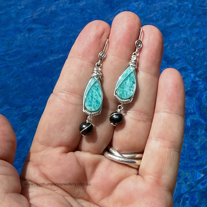 Turquoise amazonite gemstones and black tahitian pearl earrings held in a hand against a blue water background
