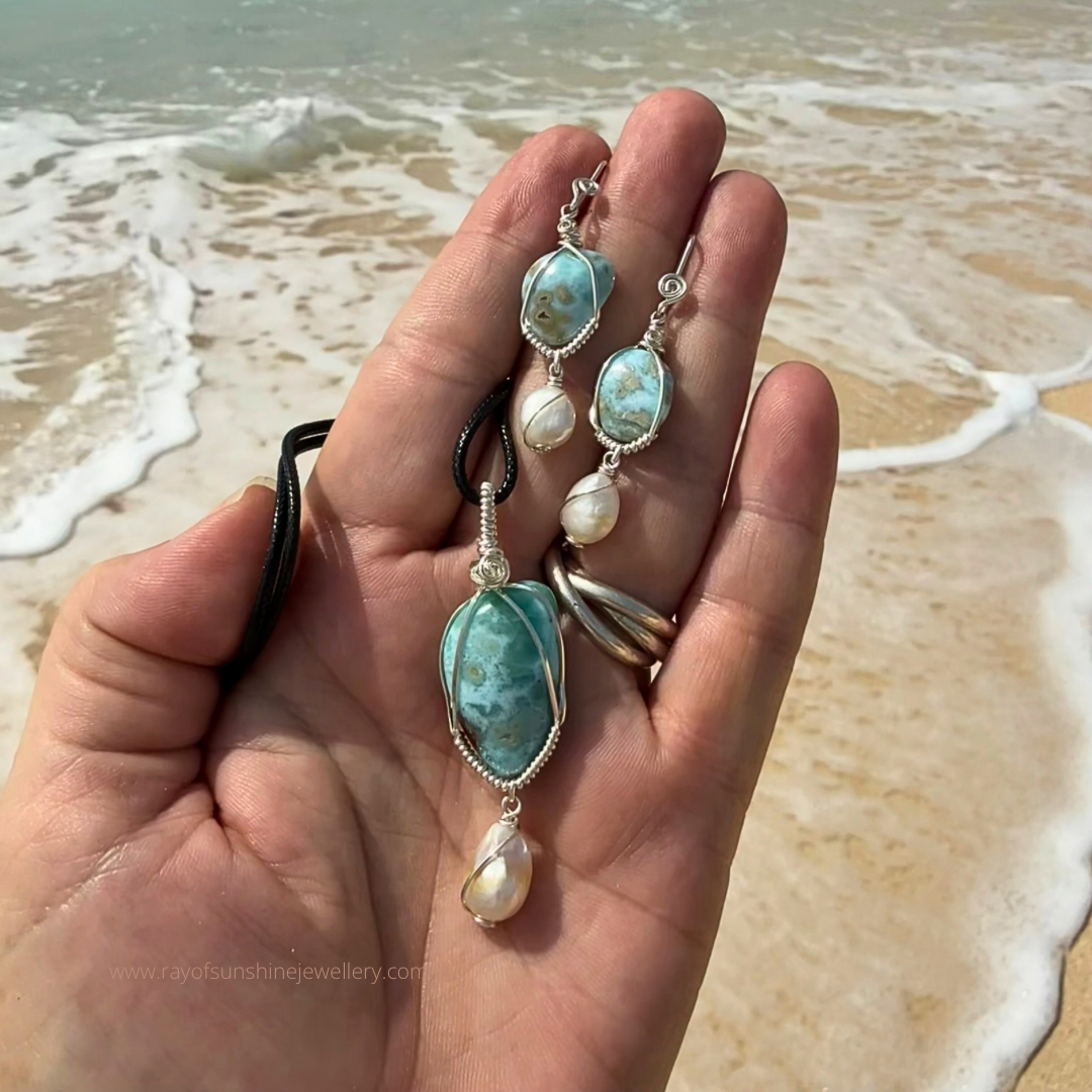 Blue green larimar and pearl earrings held in a hand with ocean waves in the background