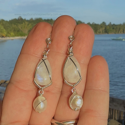 Teardrop-shaped blue moonstone and pearl earrings with silver accents held against a scenic background.