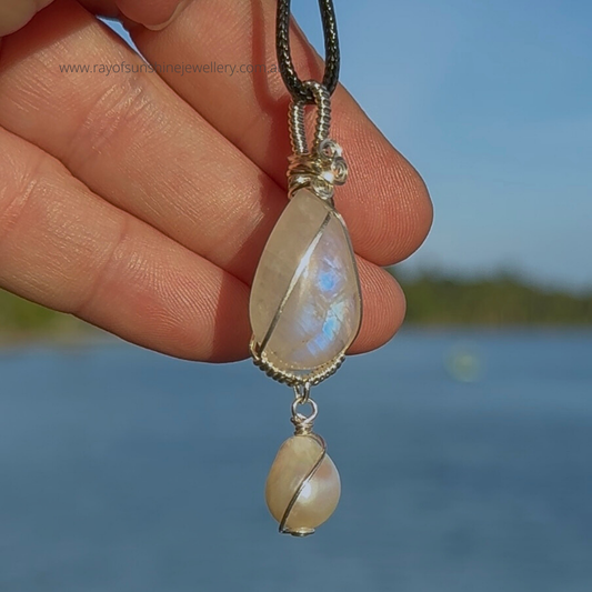Flashy blue moonstone teardrop gemstone pendant, with a white pearl wrapped in silver wire held by a hand against a blurred natural ocean background