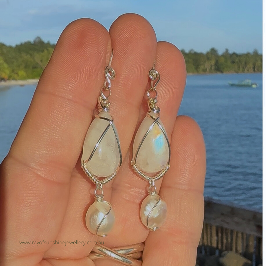 Teardrop-shaped moonstone and pearl earrings held in a hand with a scenic background of water and trees in the afternoon sunshine