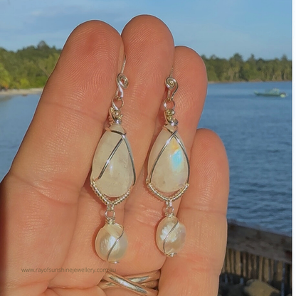 Teardrop-shaped moonstone and pearl earrings held in a hand with a scenic background of water and trees in the afternoon sunshine