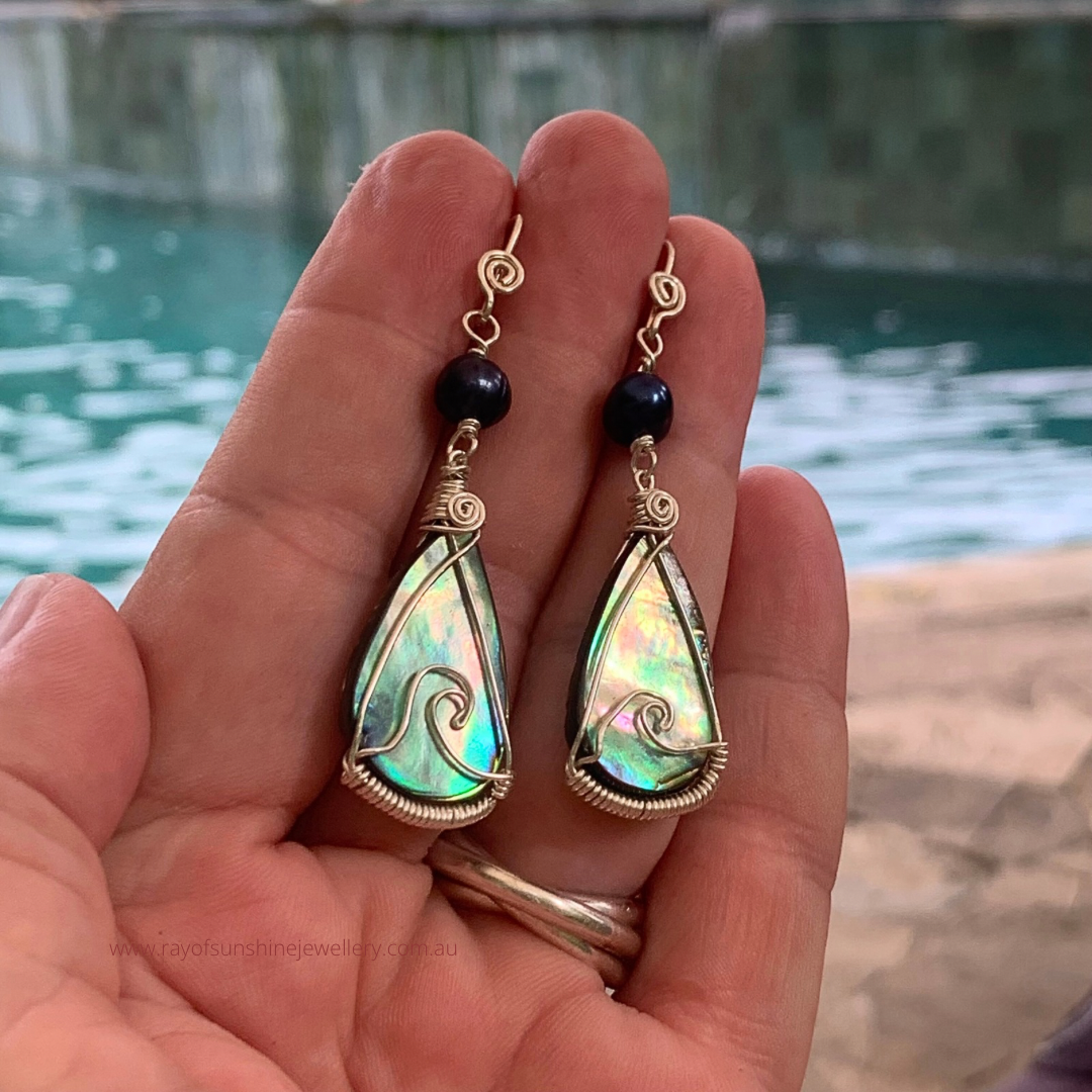 Multicoloured Teardrop-shaped albalone earrings with silver wire wrapping held in a hand overlooking water.