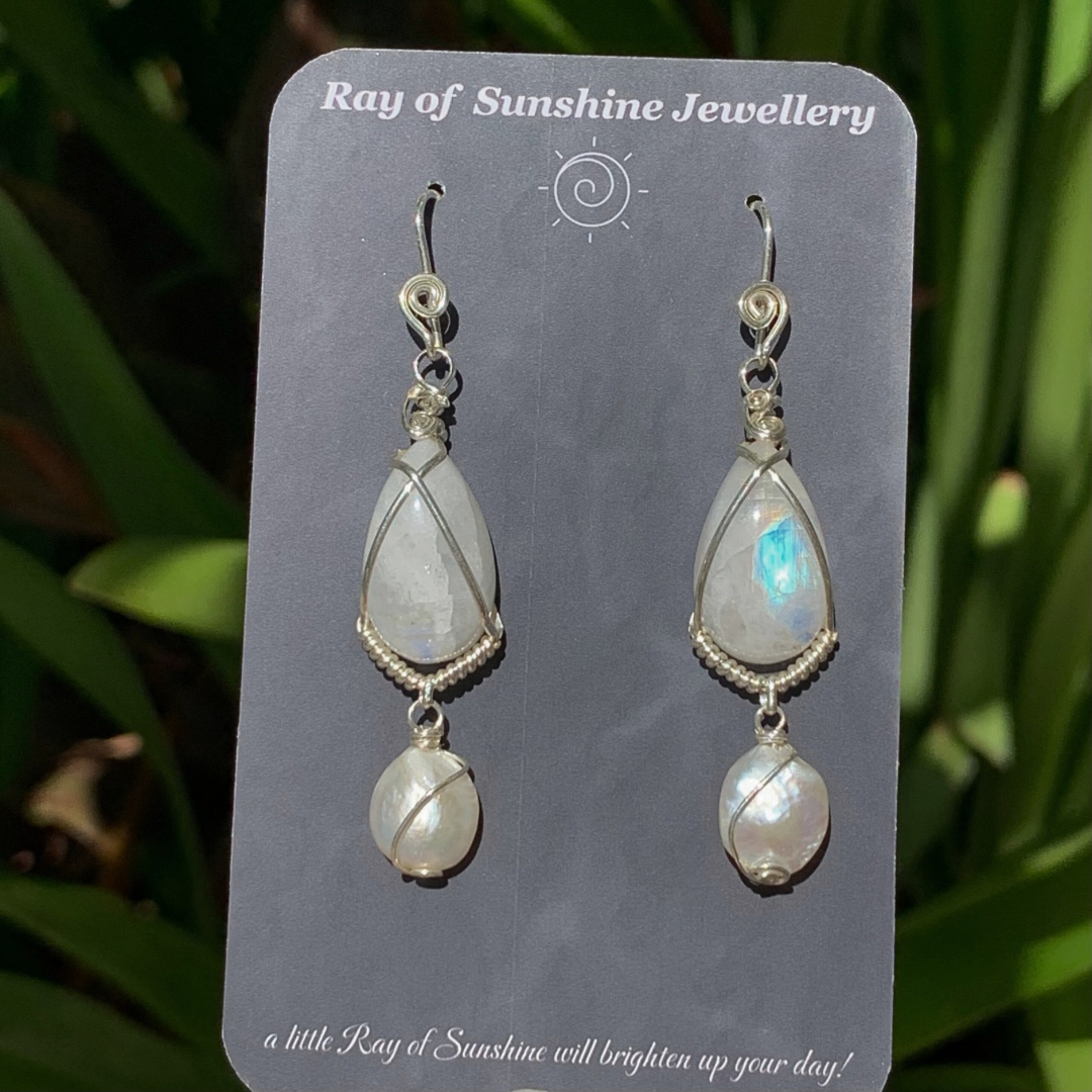 Pair of blue moonstone and pearl earrings wrapped in silver wire on a ‘Ray of Sunshine Jewellery' card against a green leafy background.