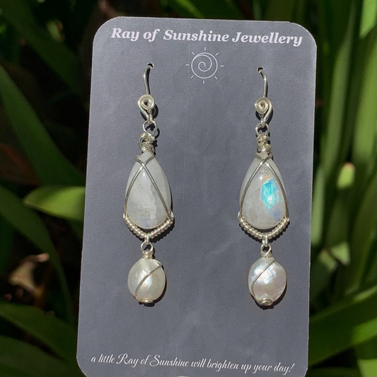 Pair of blue moonstone and pearl earrings wrapped in silver wire on a ‘Ray of Sunshine Jewellery' card against a green leafy background.