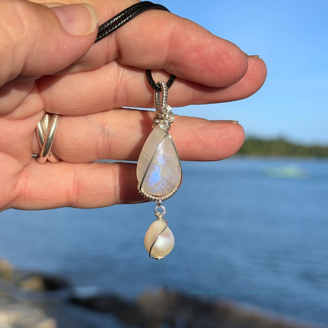 Hand holding a blue moonstone gemstone and white pearl necklace against a blurred ocean background