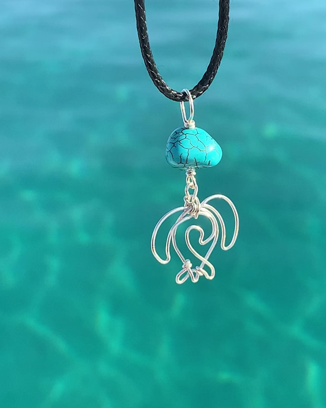 Ray of Sunshine Jewellery- my hand made Sea Turtles are hanging from a beautiful Turquoise Howlite bead and a black cord hanging over crystal clear turquoise coloured water in the sunshine. just stunning